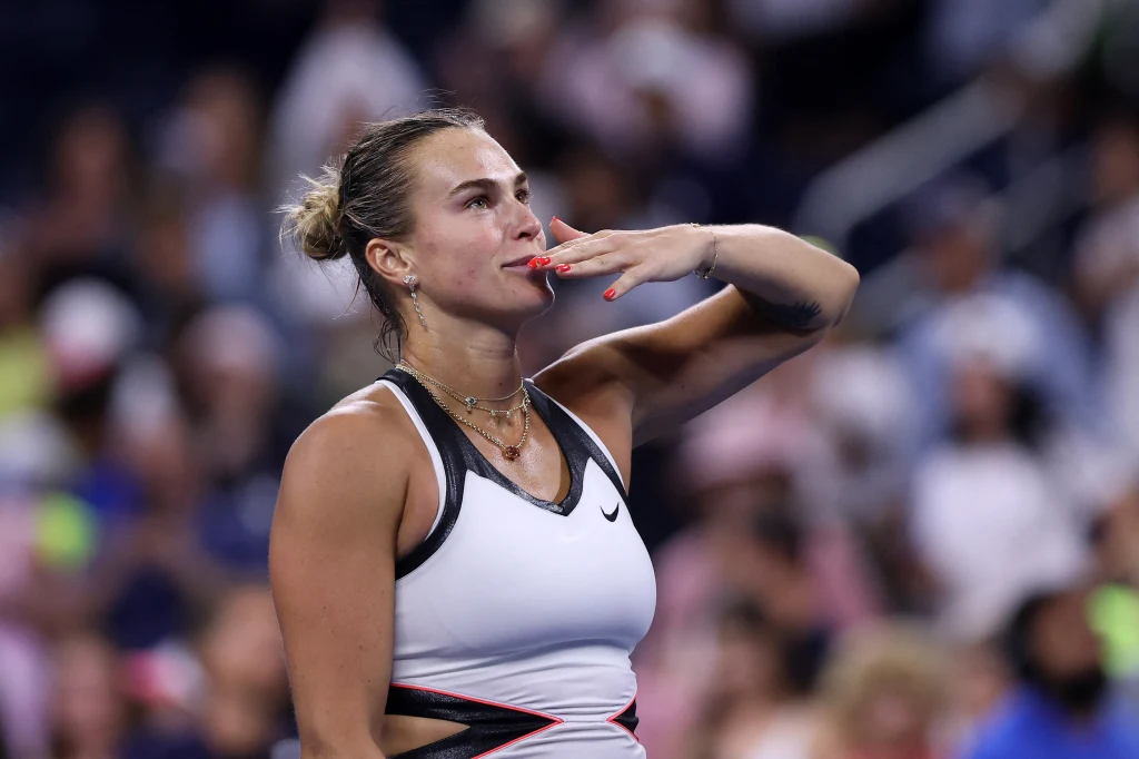 Aryna Sabalenka Tenisistka ubrana w sportowy strój wykonuje gest pocałunku w stronę publiczności na korcie, w tle widoczni są rozmyci kibice.