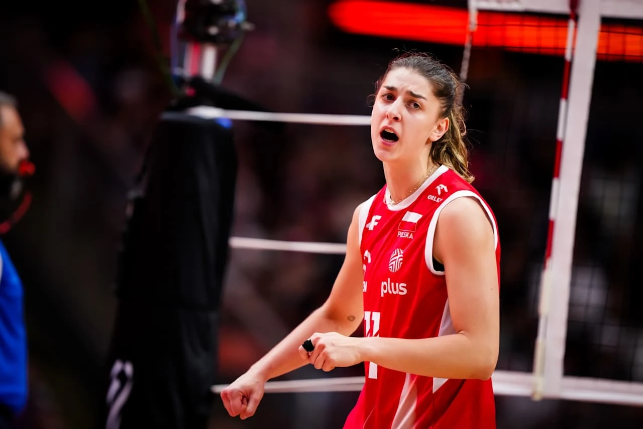 Zawodniczka drużyny siatkarskiej w czerwonym stroju, wyrażająca emocje w trakcie meczu, na tle siatki i hali sportowej, skupienie na wyrazie twarzy i atmosferze rywalizacji