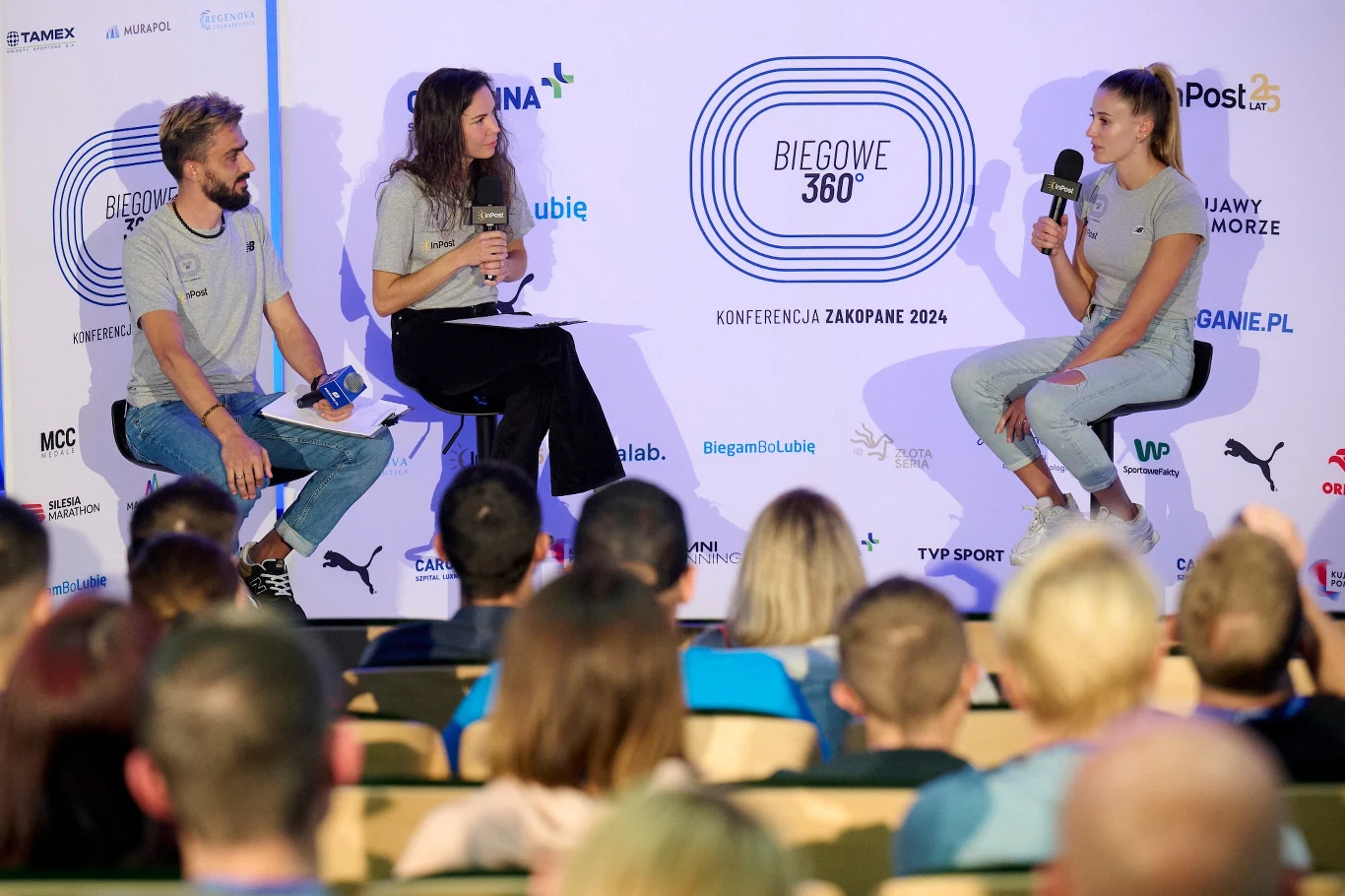 Trzech prelegentów siedzi na scenie podczas konferencji Biegowe 360°, rozmawiając ze sobą z mikrofonami w ręku, tło stanowią logotypy sponsorów oraz nazwa konferencji, publiczność siedzi odwrócona tyłem na krzesłach.