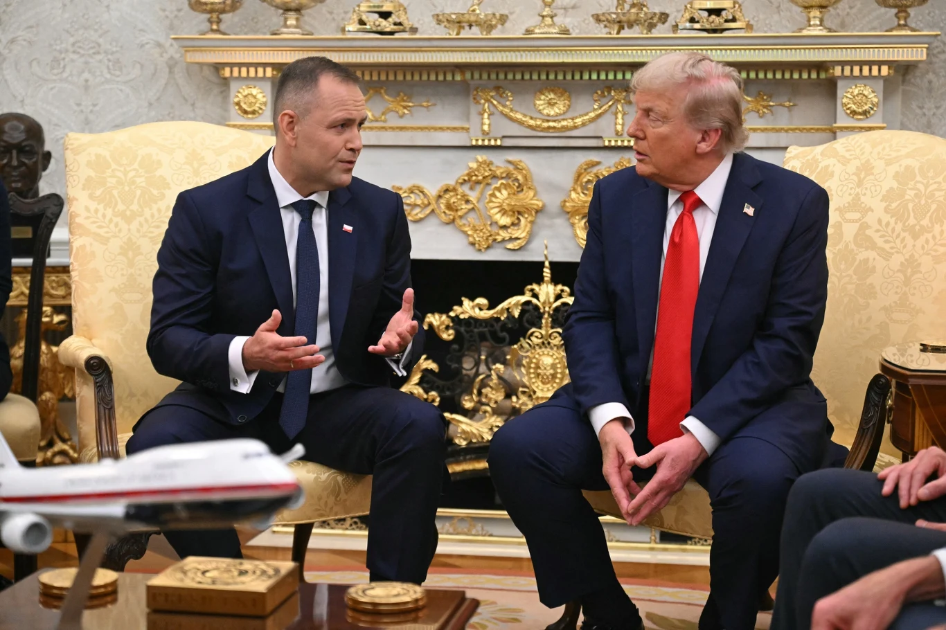 Dwóch mężczyzn w garniturach rozmawia ze sobą siedząc na eleganckich fotelach w bogato zdobionym pomieszczeniu, między nimi znajduje się kominek oraz ozdobne elementy. Na stoliku widoczny jest model samolotu.