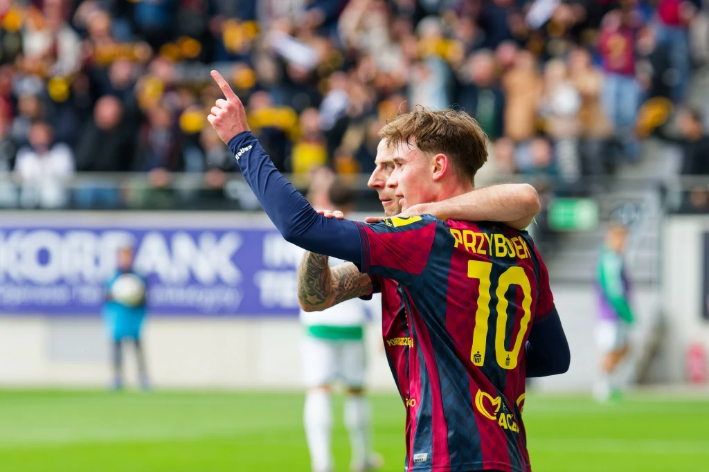 Dwóch piłkarzy ubranych w stroje meczowe obejmuje się na murawie stadionu, jeden z zawodników z widocznym nazwiskiem i numerem 10 unosi rękę, w tle kibice na trybunach.