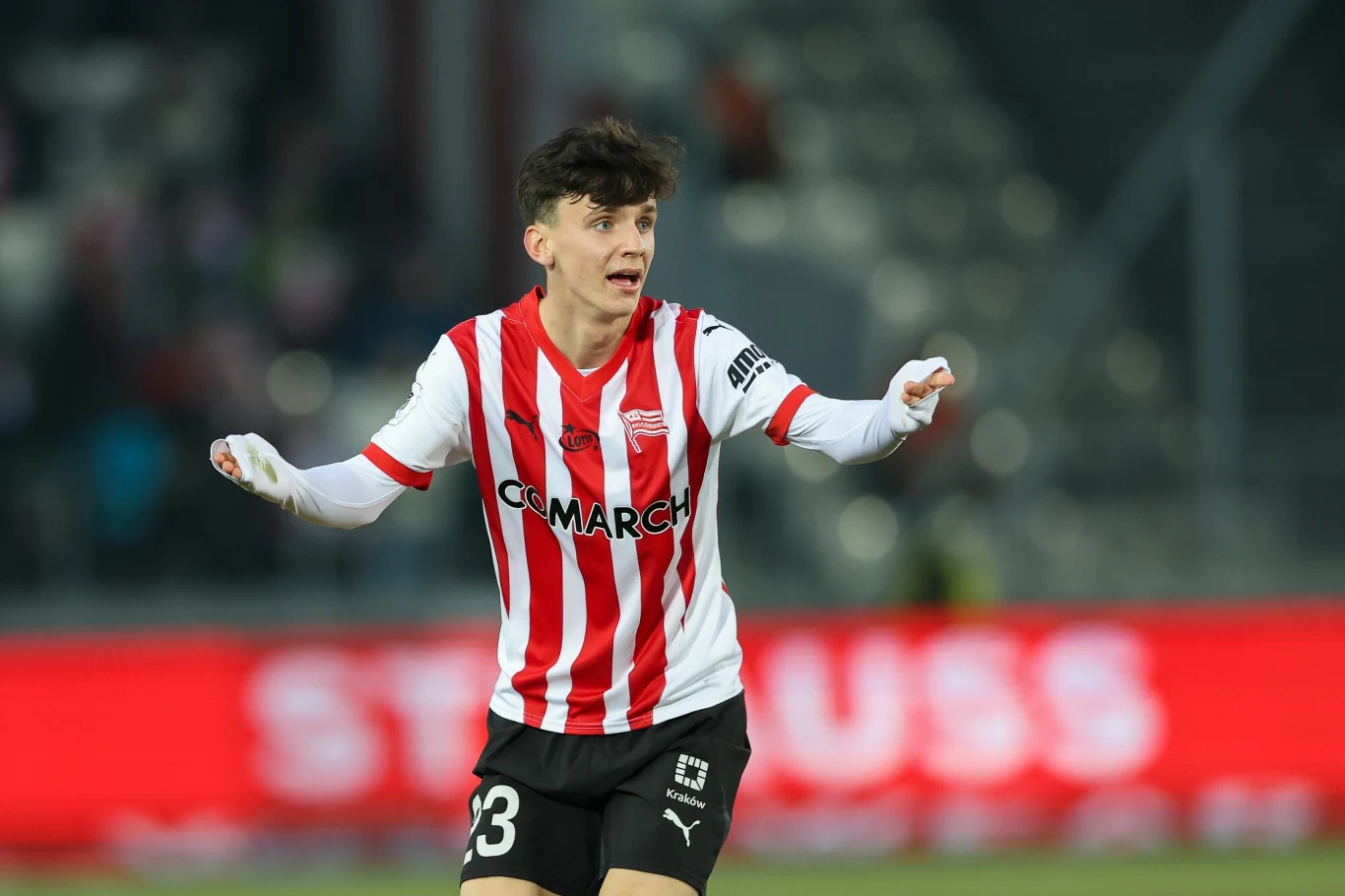 Piłkarz w biało-czerwonej koszulce z numerem 23 gestykuluje podczas meczu na stadionie, w tle rozmazane trybuny i neonowy baner reklamowy.