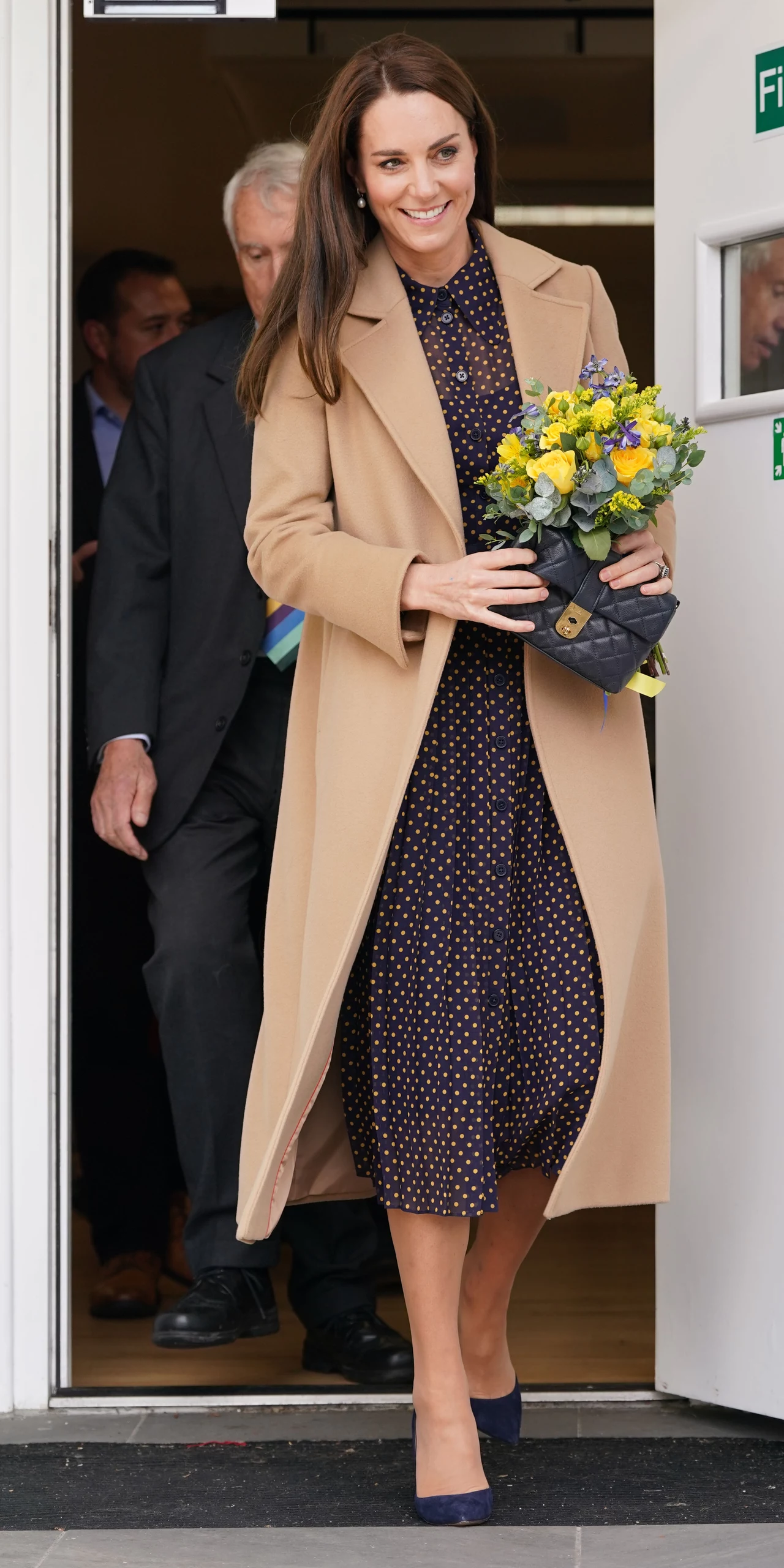 Księżna Kate uwielbia brązy jesienią z dodatkiem kropek na sukience Elegancko ubrana kobieta z długimi włosami wychodzi z budynku, trzyma bukiet żółtych kwiatów oraz czarną torebkę, ubrana w beżowy płaszcz i granatową sukienkę w drobne kropki, za nią widoczny starszy mężczyzna.
