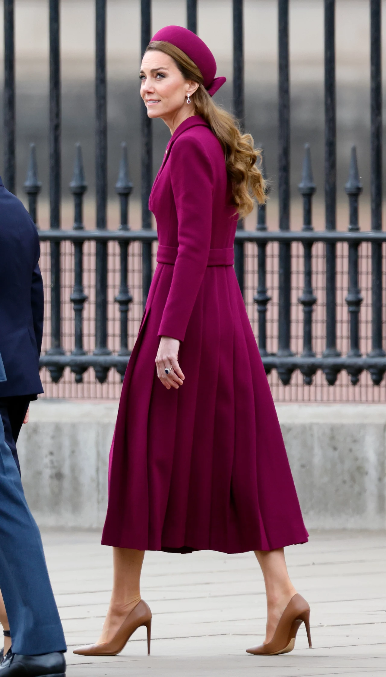 Stylizacja Kate w odcieniu fuksji to idealny pomysł na jesień Elegancka kobieta w długim, burgundowym płaszczu i dopasowanym kapeluszu idąca chodnikiem, w tle metalowe ogrodzenie; beżowe szpilki dopełniają stylizację.