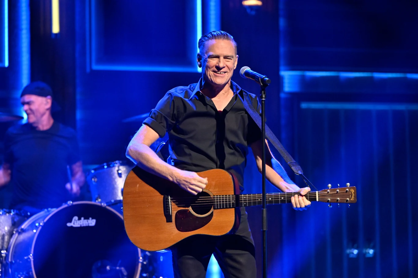 Bryan Adams wydał album we własnej wytwórni Mężczyzna grający na gitarze akustycznej na scenie, śpiewający do mikrofonu, w tle perkusista i niebieskie światła sceniczne.