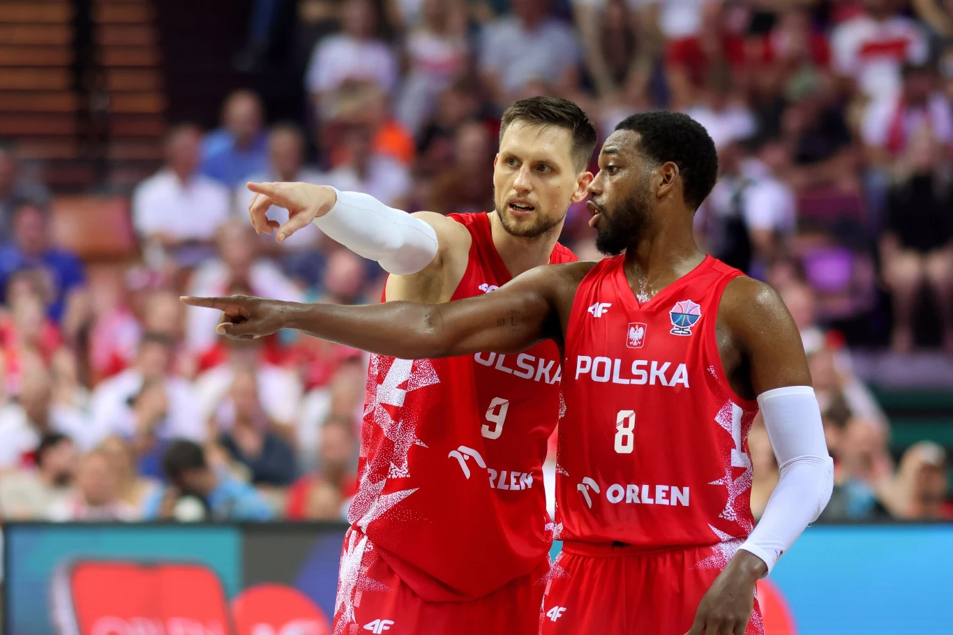 Mateusz Ponitka i Jordan Loyd Dwóch koszykarzy reprezentacji Polski w czerwonych strojach sportowych gestykuluje i patrzy w tym samym kierunku podczas meczu, w tle widownia na trybunach.