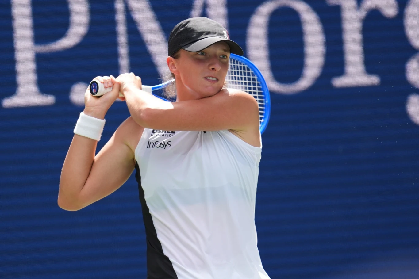 Tenisistka w białej koszulce i czapce z daszkiem przygotowuje się do wykonania uderzenia rakietą tenisową na tle niebieskiego kortu z fragmentem napisu w tle.