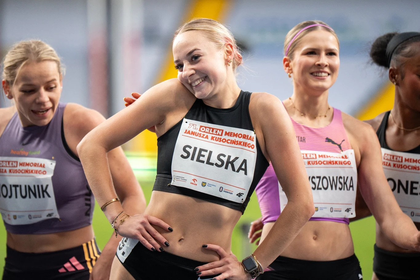 Alicja Sielska Zawodniczki lekkoatletyczne ubrane w stroje sportowe z numerami startowymi, stojące obok siebie na hali, uśmiechnięte i zrelaksowane przed lub po starcie podczas zawodów lekkoatletycznych.