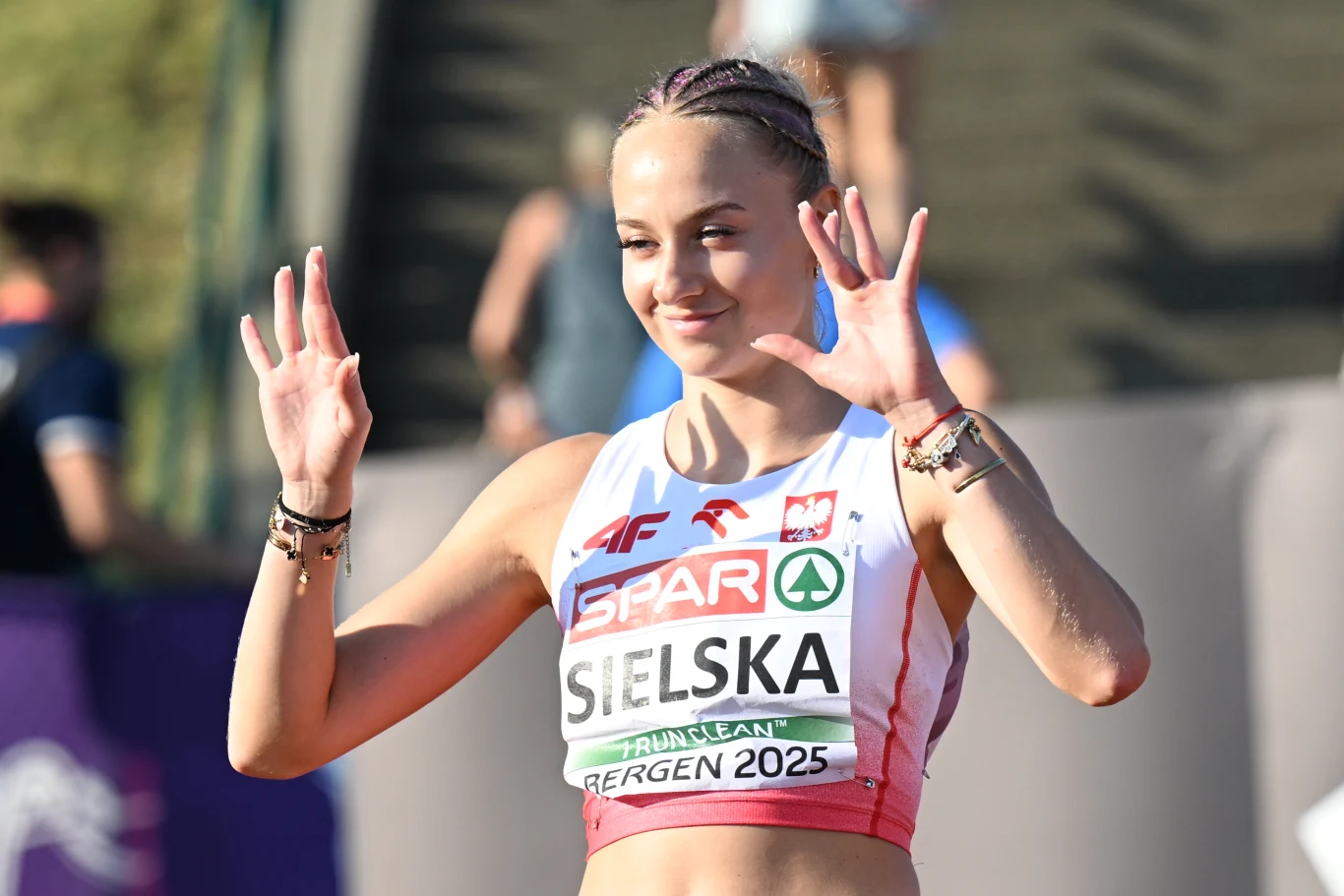 Alicja Sielska Uśmiechnięta lekkoatletka w stroju sportowym z numerem startowym, unosząca obie dłonie w górę, w tle rozmazane sylwetki innych osób i bieżnia.