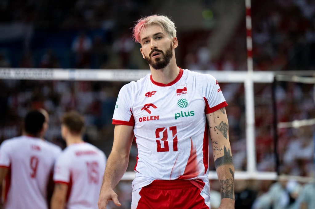 Tomasz Fornal Siatkarz w biało-czerwonym stroju z numerem 81, z tatuażami na rękach, stoi na boisku; w tle widoczni inni zawodnicy drużyny ubrani w podobne stroje oraz siatka.