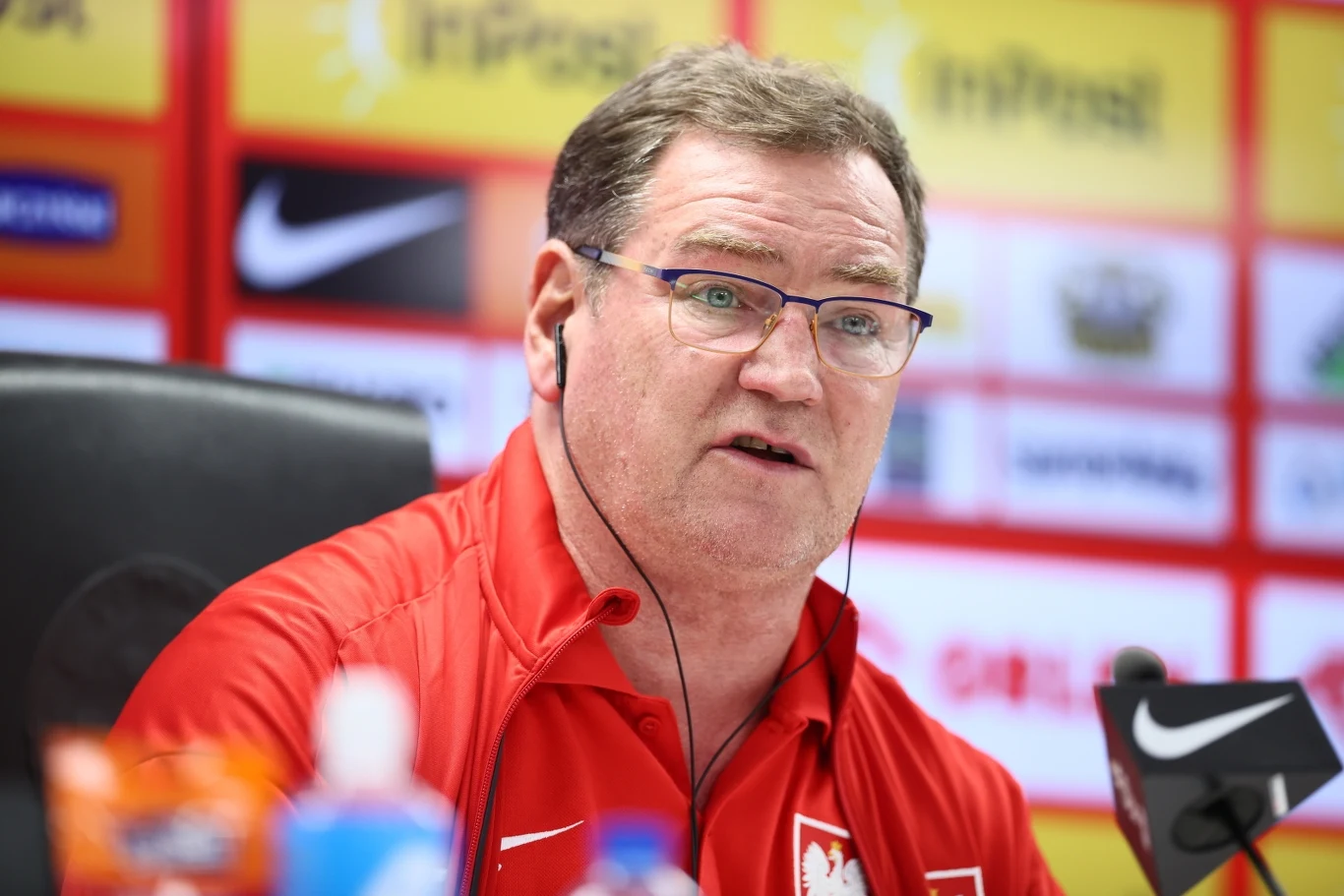 Mężczyzna w czerwonej sportowej koszuli z logo reprezentacji Polski udziela wypowiedzi podczas konferencji prasowej, w tle widoczne są logotypy sponsorów oraz tło związane z wydarzeniem sportowym.