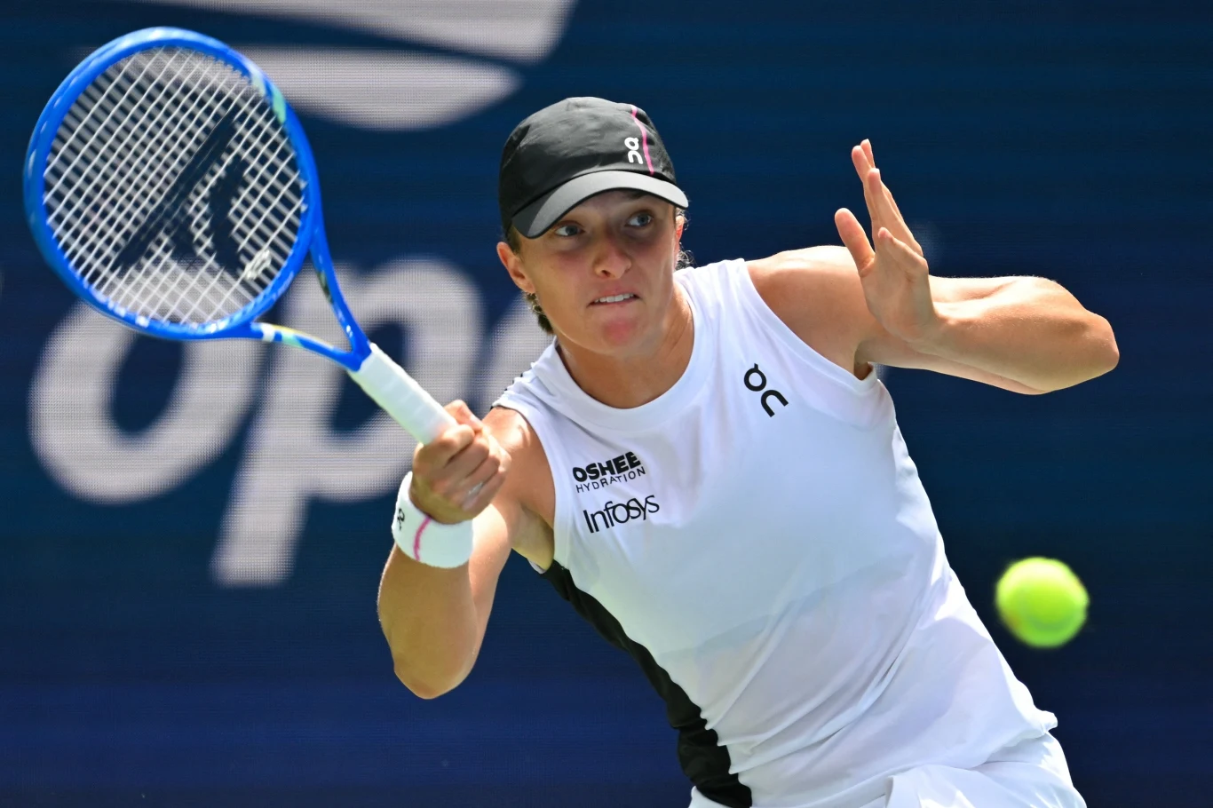 Zawodniczka tenisowa w białym stroju z niebieską rakietą tenisową w trakcie gry, skupiona na piłce lecącej z prawej strony ujęcia, na tle niebieskiego kortu