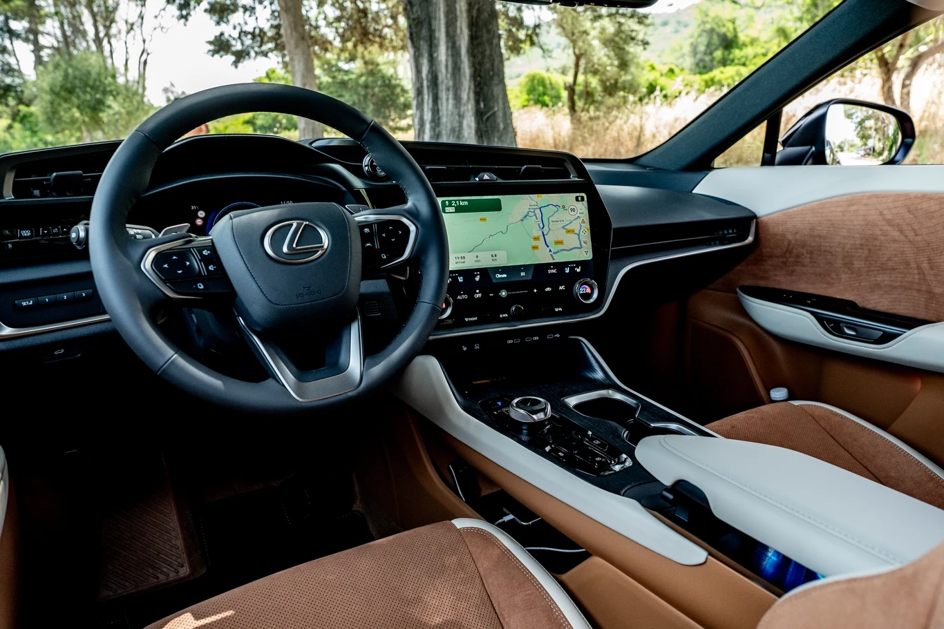 Wnętrze luksusowego samochodu marki Lexus z widoczną nowoczesną deską rozdzielczą, dużym ekranem dotykowym z nawigacją, skórzanymi siedzeniami w kolorze brązowym oraz dopracowanymi detalami wykończenia.