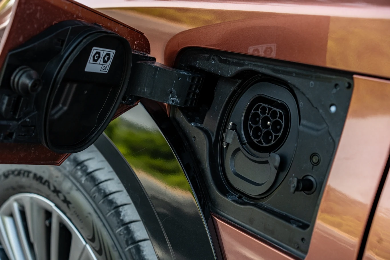 Gniazdo ładowania samochodu elektrycznego z otwartą klapką, widoczne fragmenty karoserii i tylnego koła pojazdu.