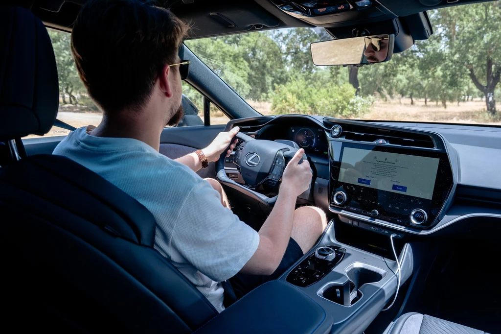 We wnętrzu debiutuje pierwszy na świecie układ steer-by-wire z wolantem, nazwany przez Japończyków One Motion Grip – bez mechanicznego połączenia kół z kierownicą. Mężczyzna z zarostem prowadzi nowoczesny samochód marki Lexus, siedząc za kierownicą, z widocznym panelem multimedialnym i komputerem pokładowym, wokół auta drzewa i krajobraz letni.