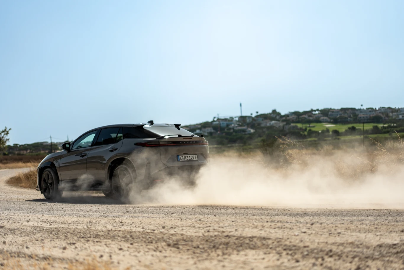 Czarny samochód osobowy jedzie po piaszczystej drodze pozostawiając za sobą chmurę pyłu, w tle widoczny krajobraz z zielonymi polami i zabudowaniami oddalonymi od drogi.