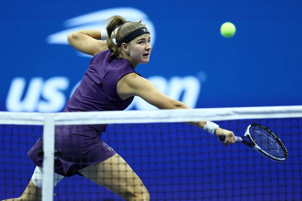 Tenisistka w fioletowym stroju i opasce na głowie dynamicznie odbija piłkę podczas meczu na kortach z niebieskim tłem i logo turnieju US Open