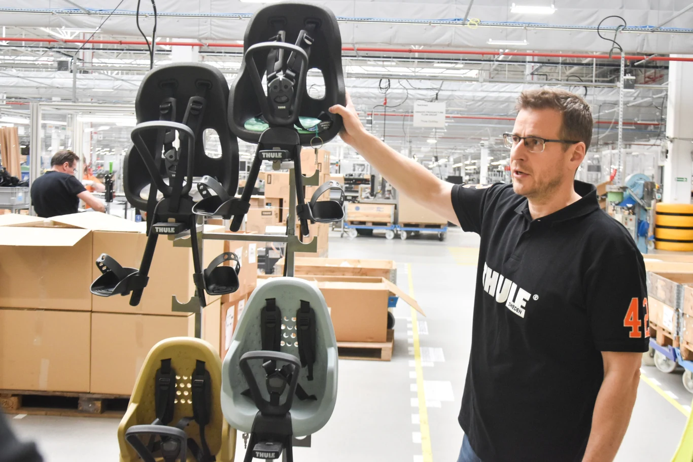 Mężczyzna w koszulce z logo firmy Thule prezentuje różne modele fotelików rowerowych dla dzieci, zawieszonych w fabrycznym otoczeniu z widoczną linią produkcyjną i kartonami w tle.