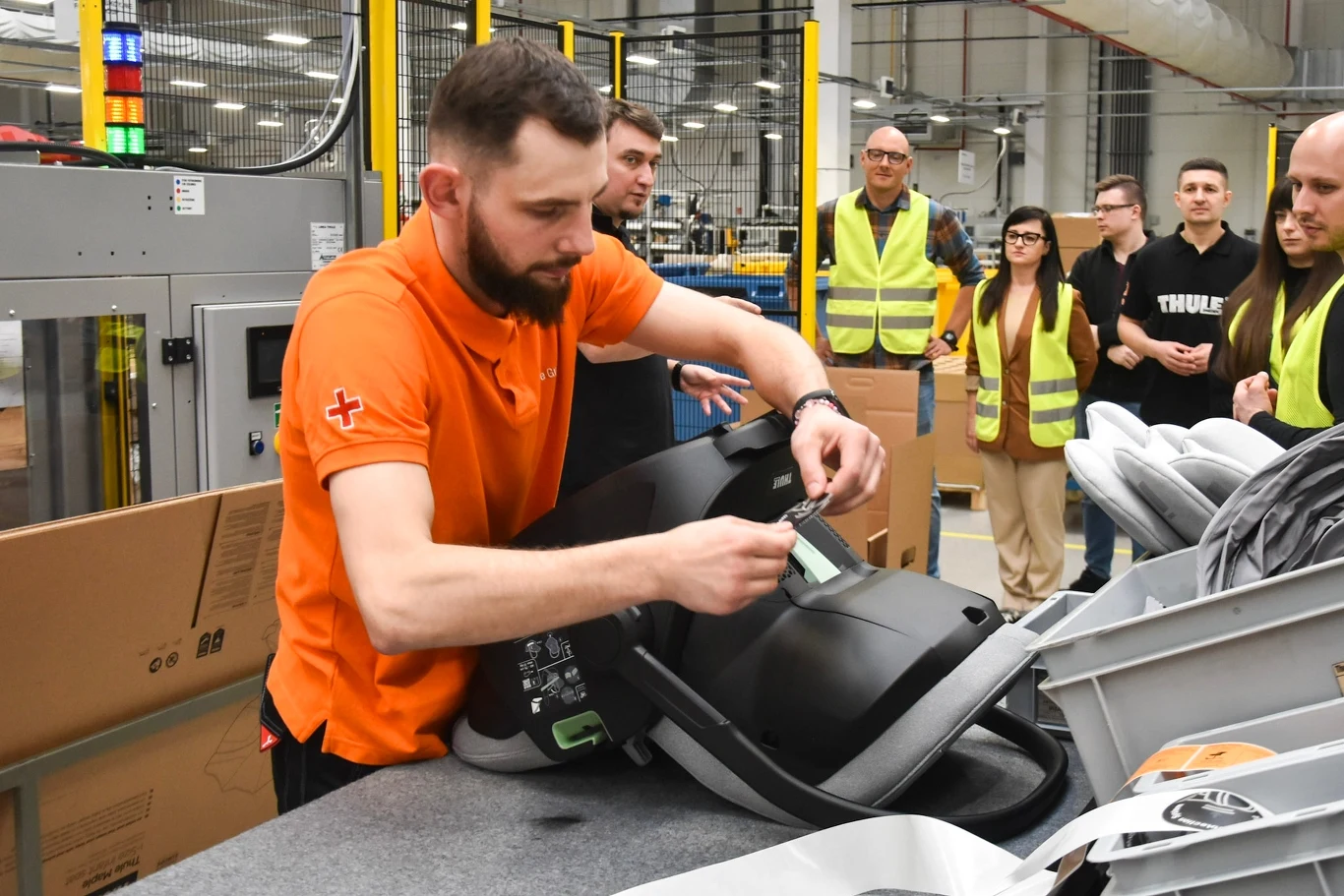 Mężczyzna w pomarańczowej koszulce sprawdza lub montuje fotelik samochodowy na stanowisku produkcyjnym, w tle grupa osób w kamizelkach odblaskowych i ubraniach roboczych obserwuje proces.