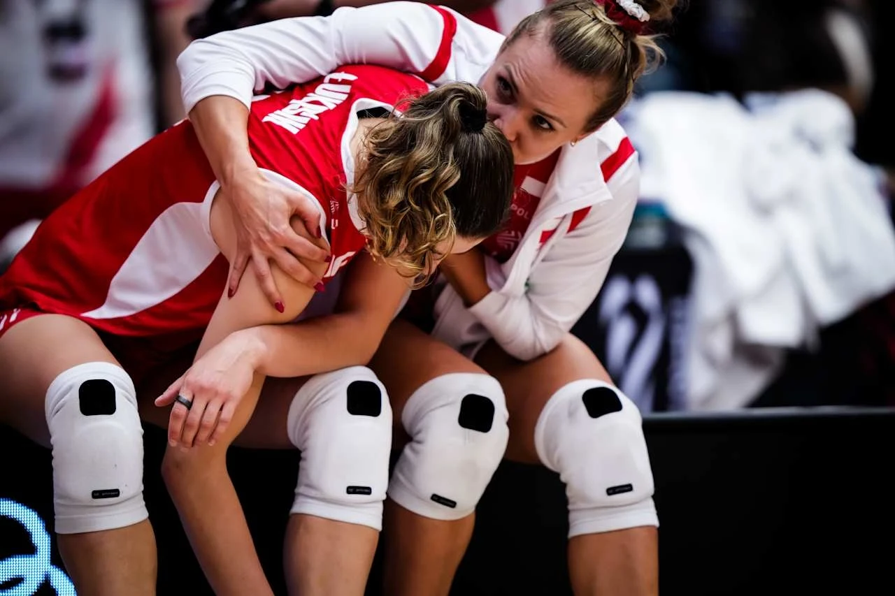 Marlena Kowalewska pocieszająca Martynę Łukasik Zawodniczka siatkówki w stroju reprezentacji Polski pocieszana przez koleżankę z drużyny, obie ubrane w stroje sportowe i ochraniacze na kolana, na ich twarzach widoczne emocje po meczu.