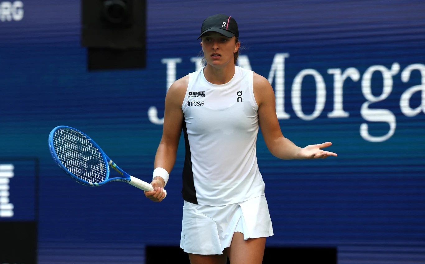 Iga Świątek Tenisistka w białej sukience sportowej i czapce, trzymająca rakietę tenisową na korcie, wyrażająca zaskoczenie lub niezadowolenie względem sytuacji na boisku. W tle widoczne logo sponsora.