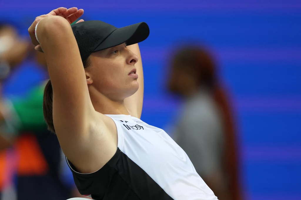Iga Świątek Sportowiec w czapce i stroju tenisowym odpoczywa, z rękami założonymi za głowę, na tle rozmytych postaci i niebieskiego boiska.