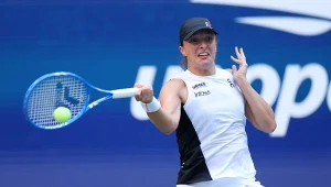 Tenisistka w białej sukience sportowej wykonuje zamach rakietą do tenisa podczas uderzenia piłki, tle widać fragment niebieskiej ściany z białymi literami.