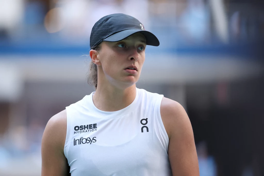 Iga Świątek Sportowa kobieta w białej koszulce, z daszkiem na głowie i logotypami sponsorów, patrząca w bok na tle boiska tenisowego.