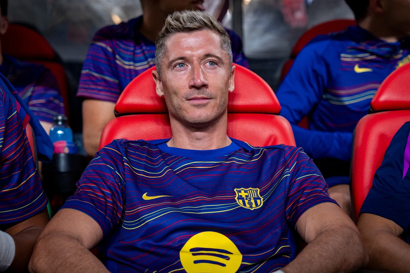 Piłkarz w koszulce FC Barcelony siedzi na czerwonym stadionowym fotelu, w tle widoczni są inni zawodnicy drużyny.