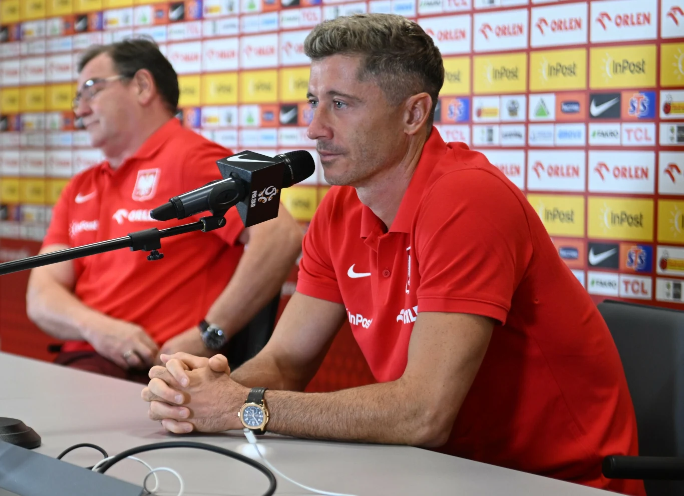 Dwóch mężczyzn w czerwonych koszulkach z logo reprezentacji Polski siedzi przy stole podczas konferencji prasowej, na tle ściany z logotypami sponsorów, jeden z nich odpowiada na pytania przez mikrofon.