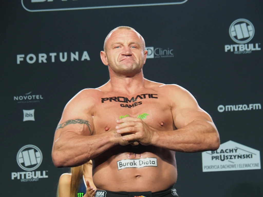 Mariusz Pudzianowski Umięśniony, łysy mężczyzna pozuje na tle ściany z logo sponsorów, z widocznymi tatuażami na ciele i oznaczeniami reklamowymi na klatce piersiowej oraz brzuchu, w pozycji zaciśniętych dłoni.