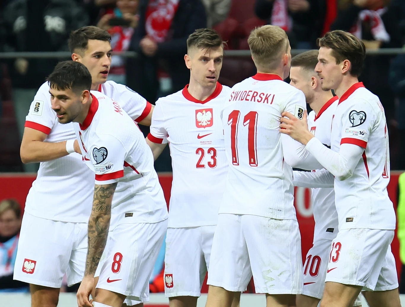 Grupa piłkarzy reprezentacji Polski w białych strojach z czerwonymi elementami stoi razem na boisku, niektórzy z nich wymieniają gesty wsparcia, w tle widać fragment trybuny z kibicami.