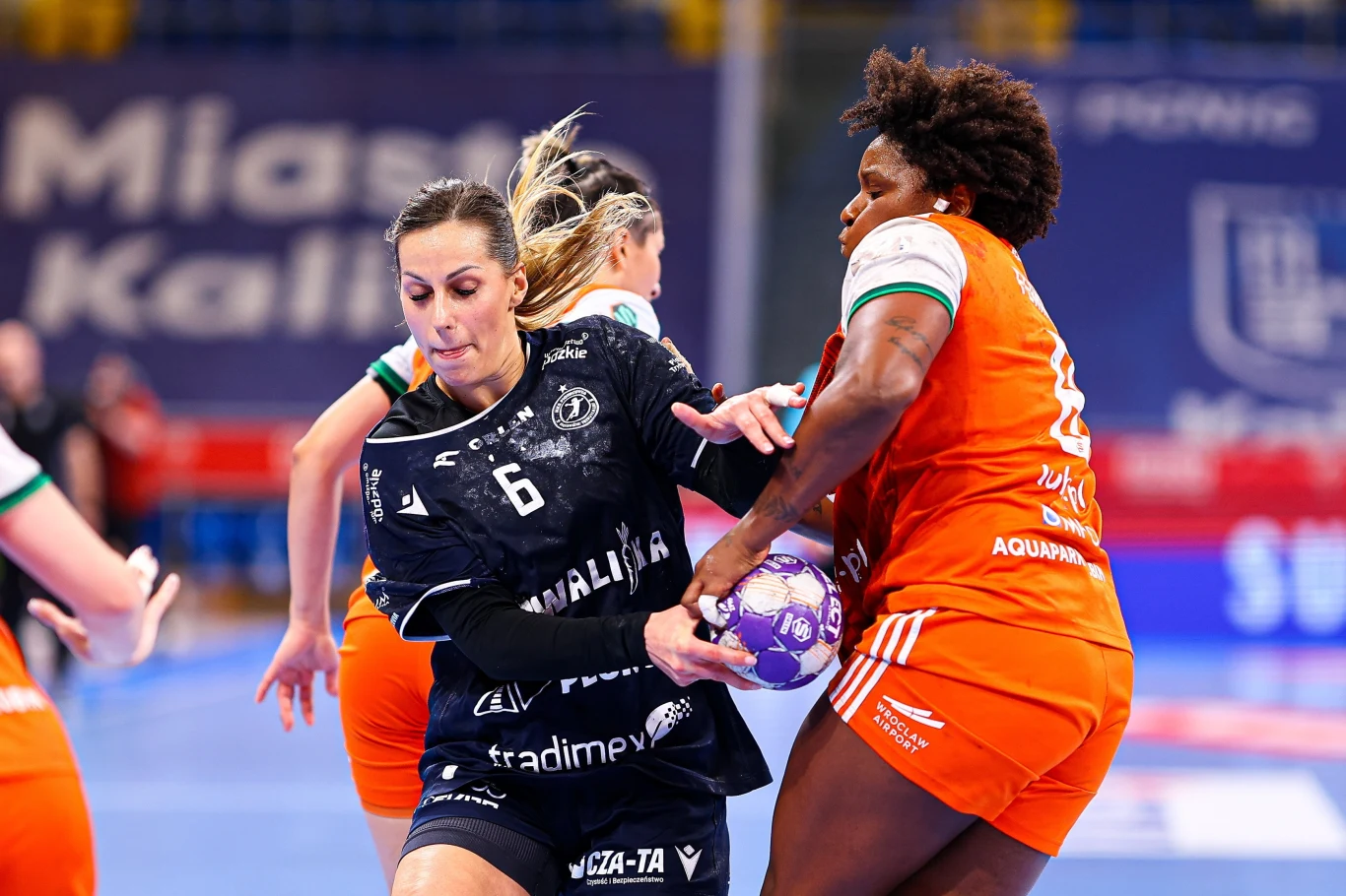 Patrycja Noga Dwie zawodniczki drużyn piłki ręcznej walczą o piłkę podczas intensywnej akcji na boisku, w tle widoczna hala sportowa oraz inne uczestniczki meczu.