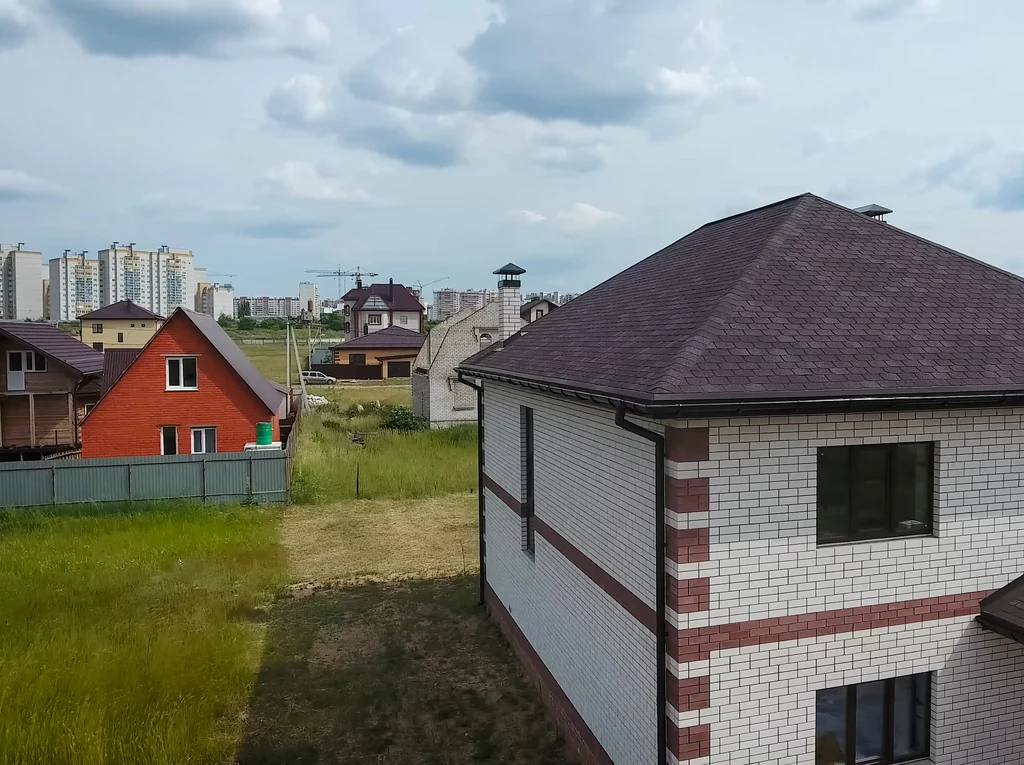 Domy jednorodzinne z cegły i drewna stojące na zielonym terenie, w tle widać osiedle wysokich bloków mieszkalnych, a nad całością rozciągają się pochmurne niebo.