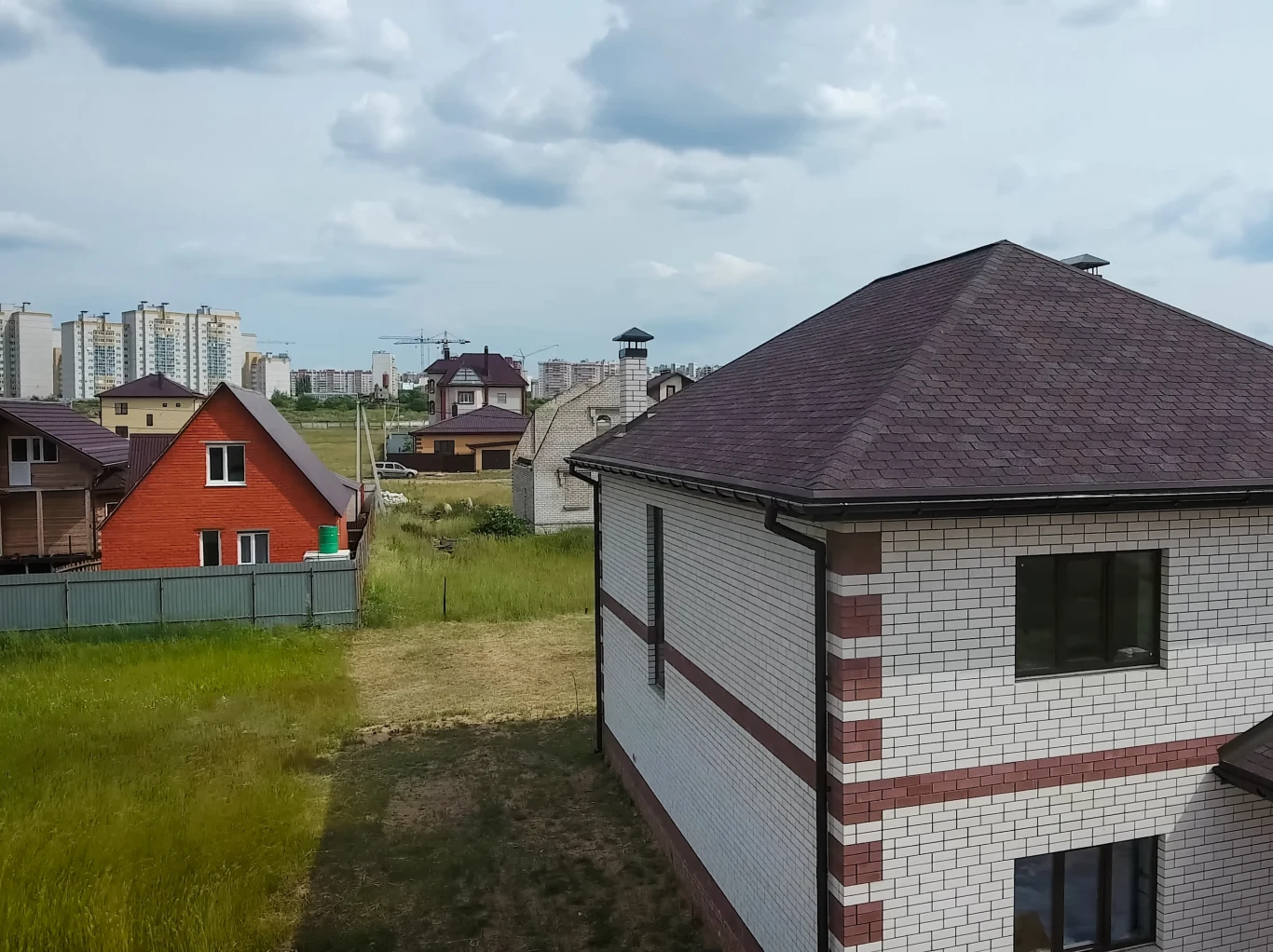 Domy jednorodzinne z cegły i drewna stojące na zielonym terenie, w tle widać osiedle wysokich bloków mieszkalnych, a nad całością rozciągają się pochmurne niebo.