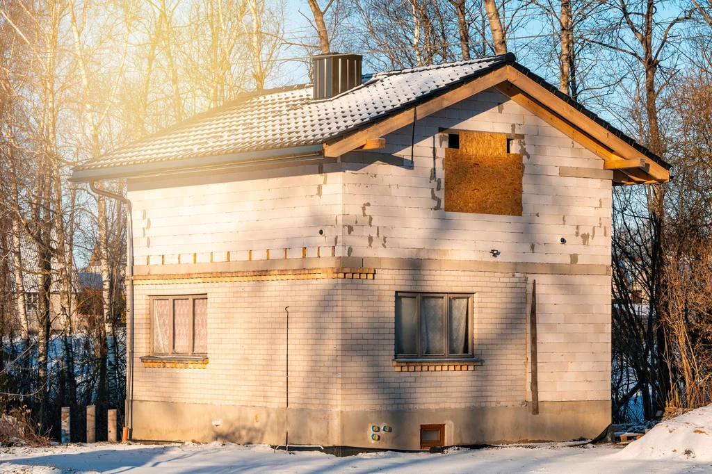 Niedokończony budynek na tle ośnieżonego krajobrazu, z oknami częściowo osłoniętymi deskami i widoczną, nieotynkowaną elewacją, otoczony przez nagie drzewa w zimowej aurze.