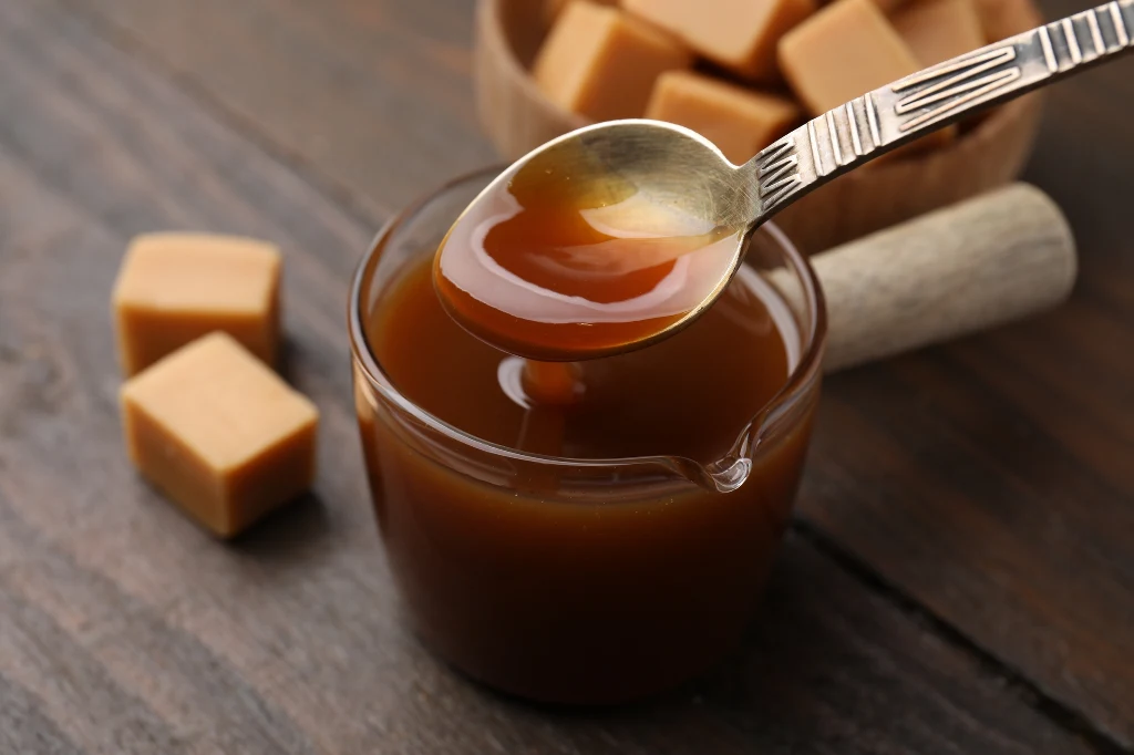 Bazą syropu a'la cola jest karmel Szklane naczynie wypełnione gęstym karmelem, nad którym unosi się łyżka z porcją płynnego karmelu, w tle kilka kostek toffi na drewnianym stole.