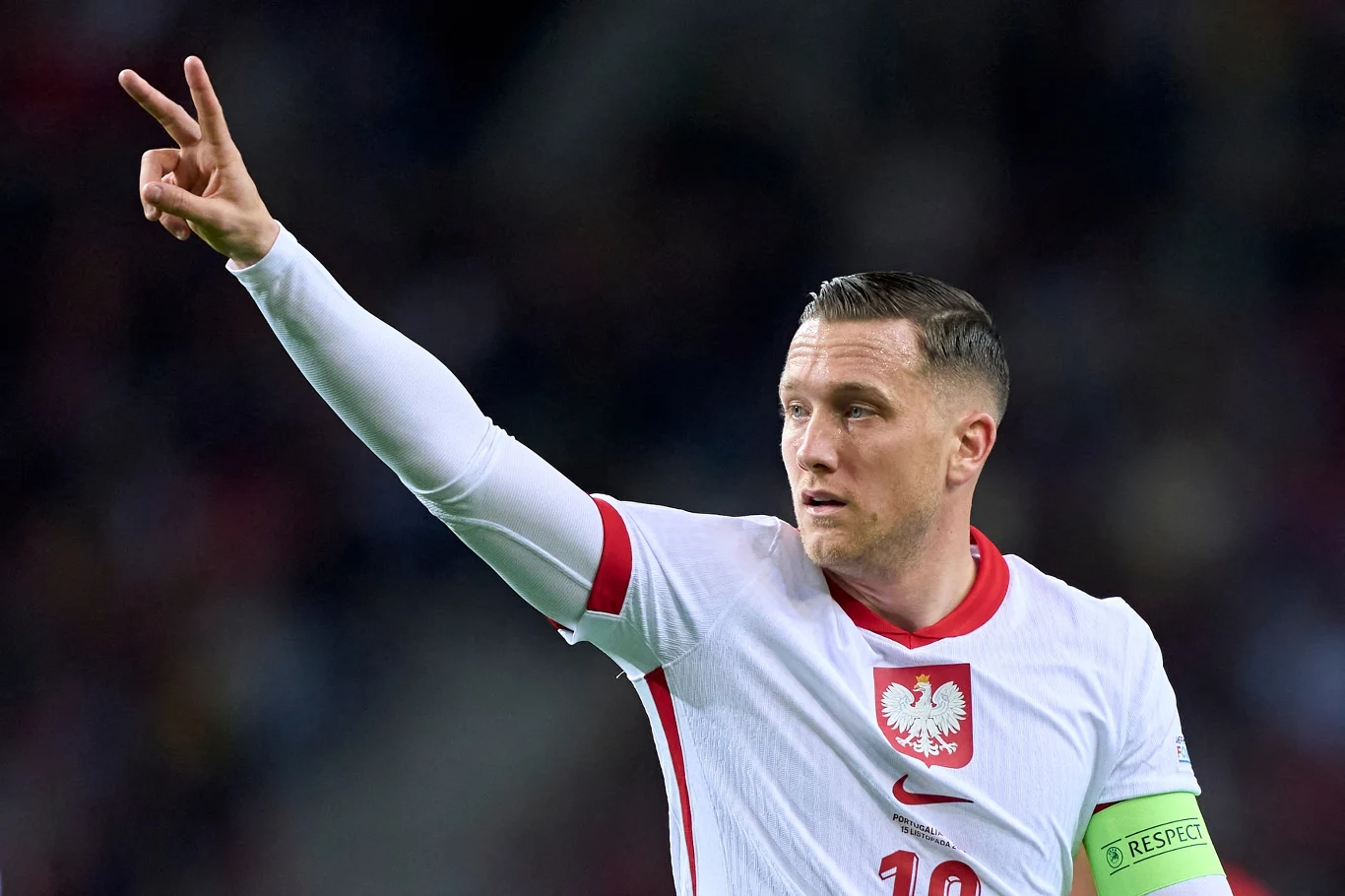 Polski piłkarz w białej koszulce z orłem na piersi i opaską kapitana na ramieniu unosi rękę, pokazując dwa palce w geście zwycięstwa.