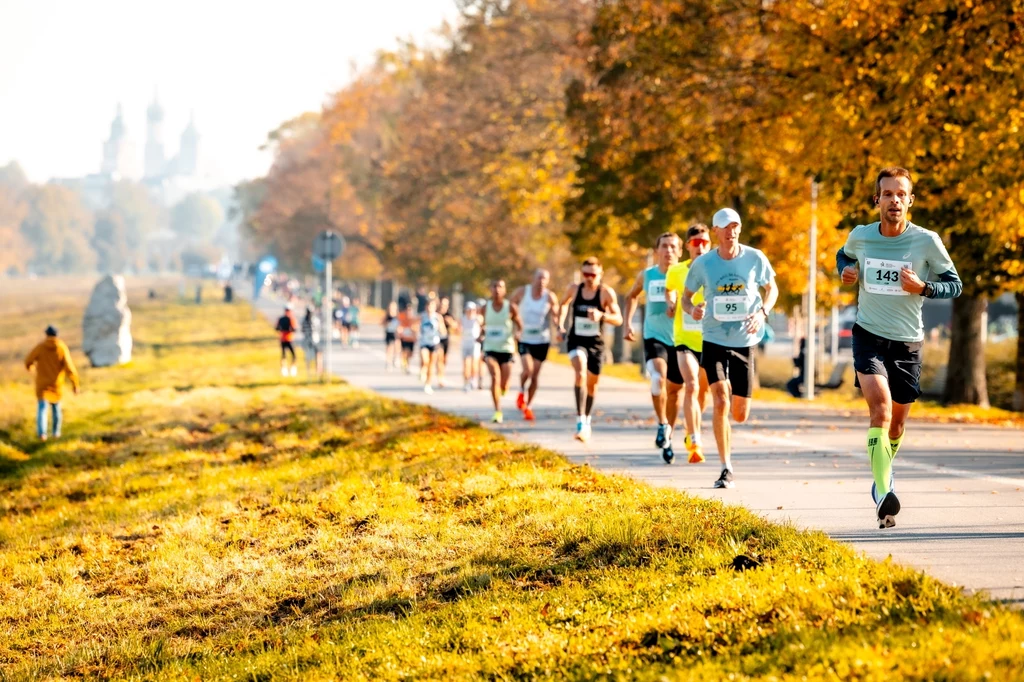 Uczestnictwo w krakowskich wydarzeniach z pozycji kibica to szansa na poczucie pulsu miasta Grupa biegaczy uczestniczących w maratonie biegnąca alejką w parku, otoczona jesiennymi drzewami i żółtymi liśćmi, w oddali widoczne są zabudowania miasta.