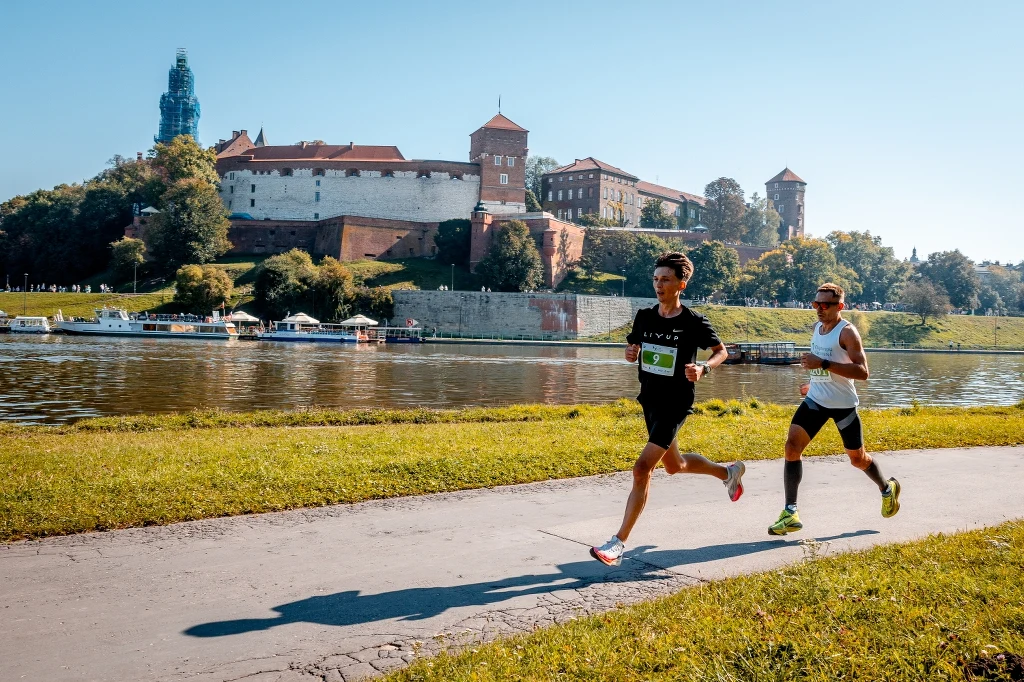 Tysiące biegaczy przybywa do Krakowa co roku, bo biegi są czymś więcej niż rywalizacją Dwóch biegaczy uczestniczących w maratonie biegnie wzdłuż Wisły, w tle panorama Wawelu z historycznym zamkiem, jasne niebo, zielone drzewa i spokojna woda podkreślają pogodną atmosferę wydarzenia sportowego.
