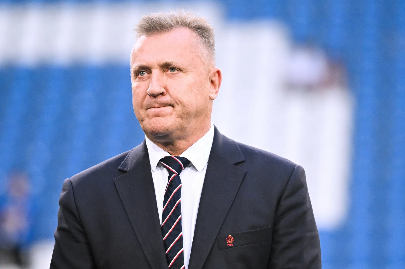 Mężczyzna w garniturze i krawacie stoi na tle niebieskich, rozmytych trybun stadionu, spoglądając lekko w bok z neutralnym wyrazem twarzy.