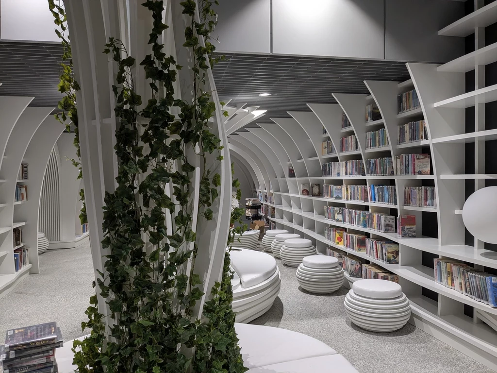 Ciekawym elementem Metroteki są meble parametryczne Nowoczesne wnętrze biblioteki z białymi regałami, okrągłymi siedziskami i zielonym pnączem oplatającym kolumnę, półki wypełnione książkami sięgają niemal sufitu, a przestrzeń sprzyja czytaniu i relaksowi.