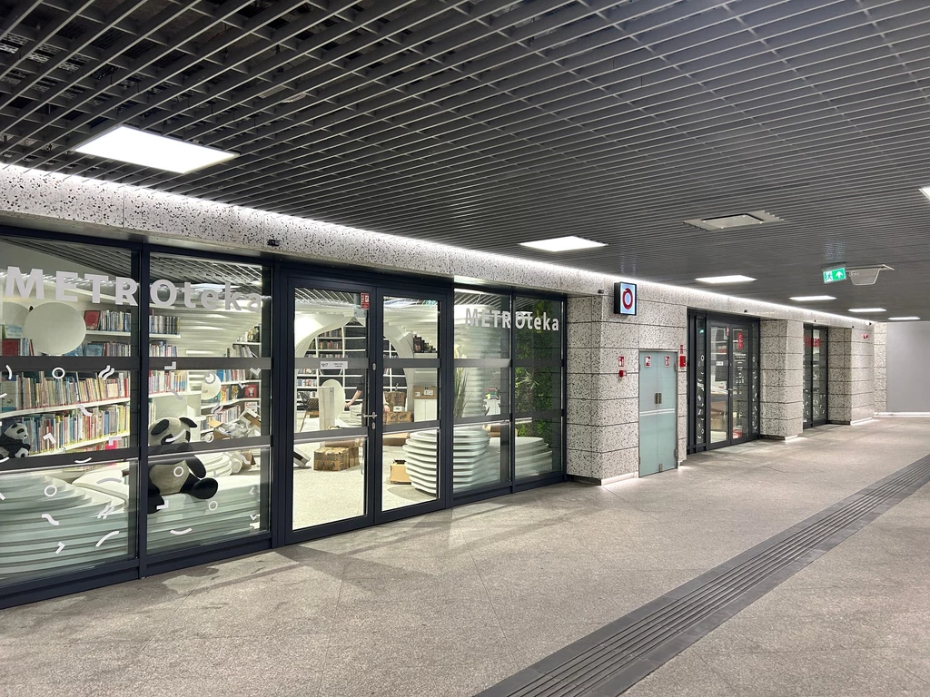 Metroteka Przeszklone drzwi prowadzące do nowoczesnej biblioteki w stacji metra, za którymi widoczne są regały z książkami oraz stoliki i krzesła do czytania, oświetlenie sufitowe tworzy jasną i przyjazną atmosferę.