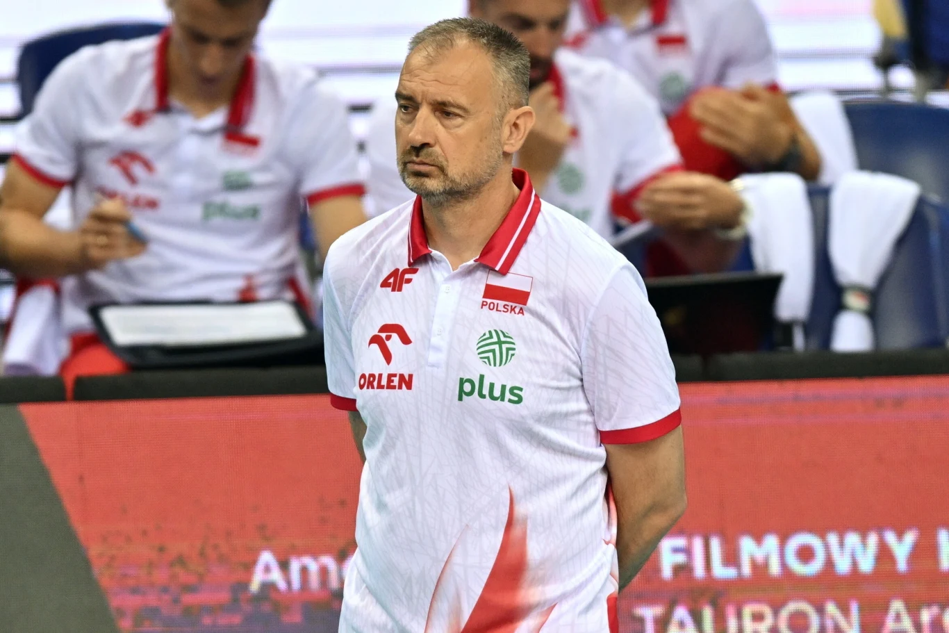 Nikola Grbić Trener drużyny siatkarskiej Polski w białej koszulce z logotypami sponsorów stoi przed ławką rezerwowych, a w tle widoczni są inni członkowie sztabu i zawodnicy.