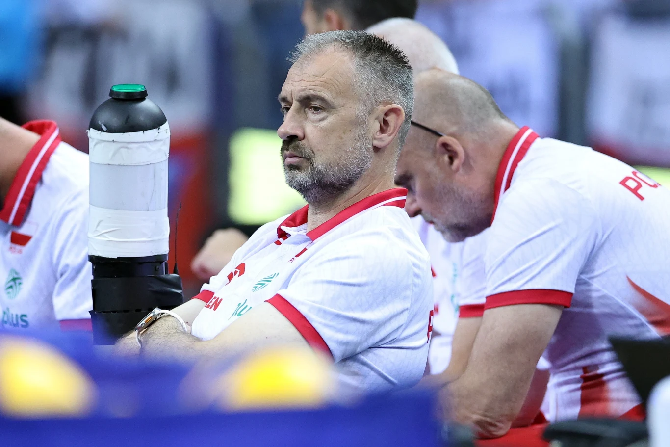 Nikola Grbić Mężczyzna w sportowej koszulce z nadrukiem flagi Polski oraz logo sponsora siedzi wśród innych członków drużyny, trzymając dużą butelkę napoju, z poważnym wyrazem twarzy, w tle rozmyte postacie.