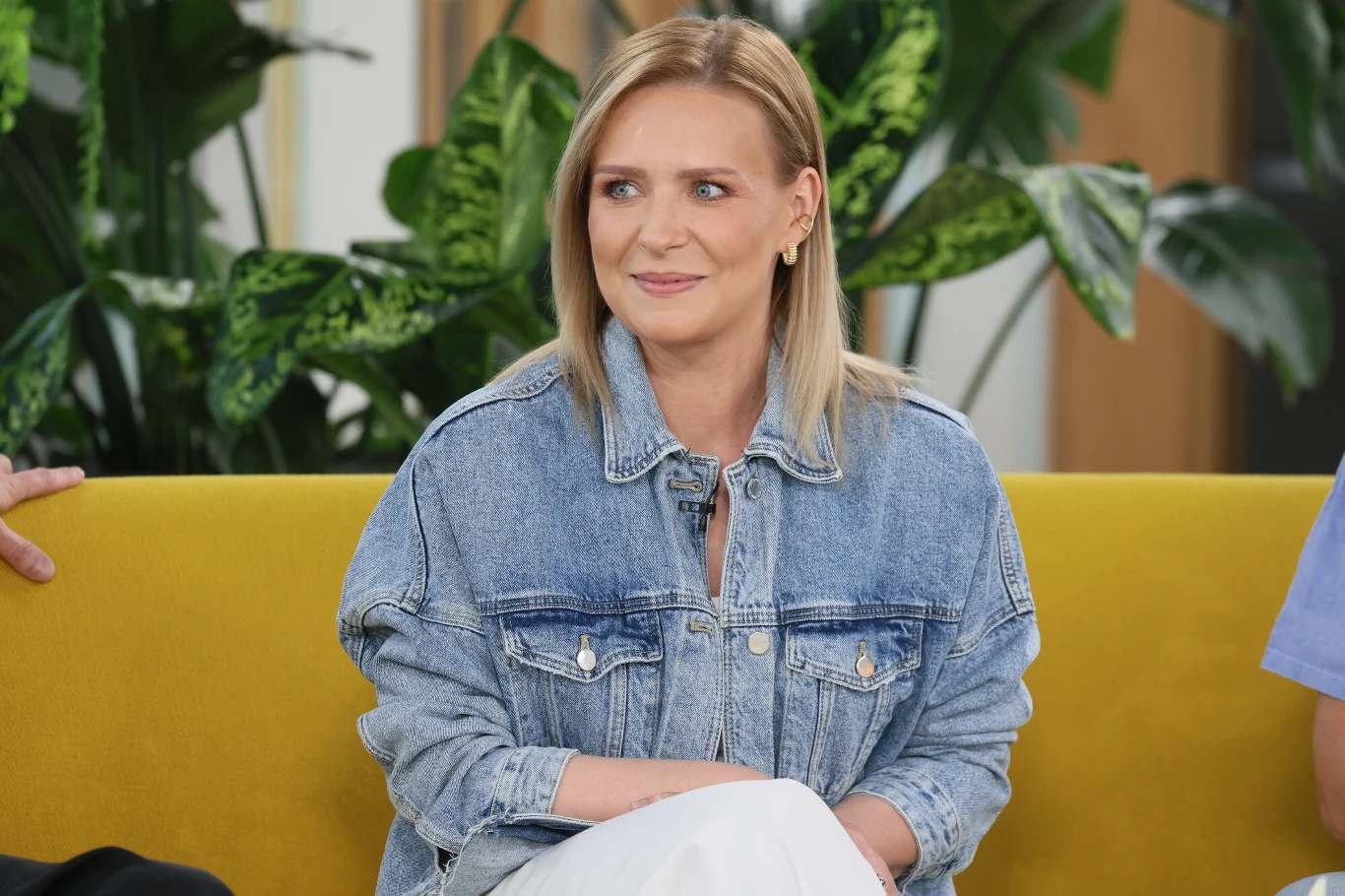 Kobieta o blond włosach siedząca na żółtej kanapie, ubrana w jasną dżinsową kurtkę, w tle widoczne zielone rośliny.