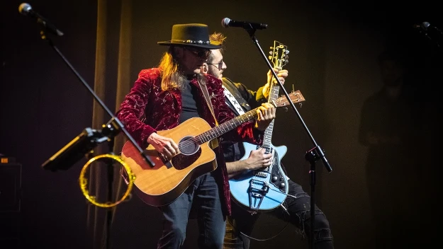 Dwóch muzyków grających na gitarach na scenie koncertowej, oświetleni światłem reflektorów, z widocznym mikrofonem i tamburynem na pierwszym planie.