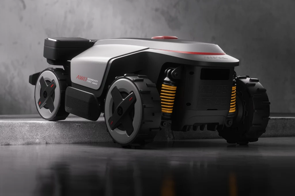 Robot koszący Roborock RockMow Z1. Nowoczesny robot ogrodowy z czterema czarnymi kołami i żółtymi amortyzatorami ustawiony na betonowej powierzchni, wykończony w czarnym i szarym kolorze z czerwonym akcentem na górze.