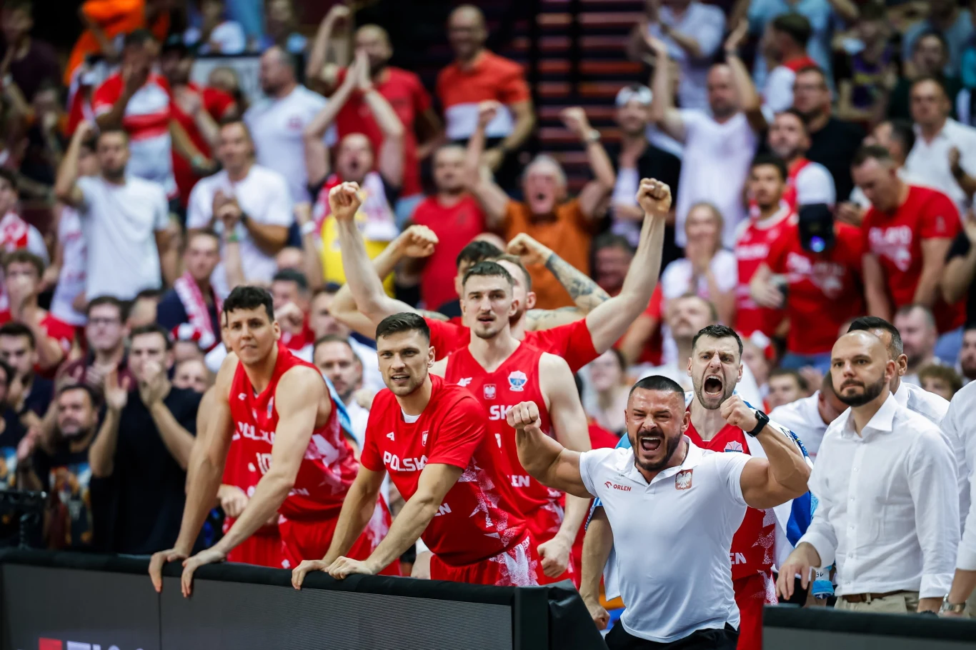 Rezerwowi reprezntacji Polski podczas mistrzostw Europy Polscy siatkarze oraz członkowie sztabu z entuzjazmem świętują zdobycie punktu podczas meczu, a tłum kibiców w tle wznosi ręce i wyraża wsparcie dla drużyny.