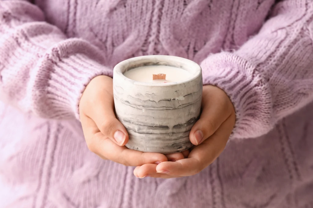 Zamiast zwykłych świec zapachowych lepiej wybrać te naturalne - rzepakowe lub sojowe Dłonie ubrane w różowy sweter trzymają szary, marmurkowy pojemnik z białą świecą i drewnianym knotem.